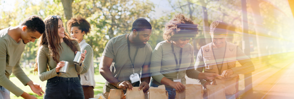 Five volunteers in a park filling brown paper bags with food items at an outdoor table under trees.