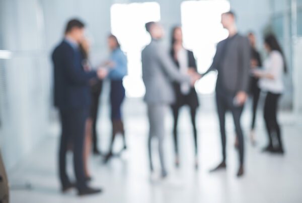 Blurred group of business professionals in suits milling about and two people shaking hands in a bright modern office lobby.