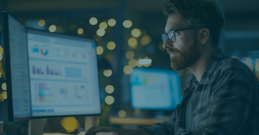 Professional reviewing Security Awareness Training analytics dashboard on dual monitors, analyzing phishing simulation results and employee security performance data in a low-light office setting.