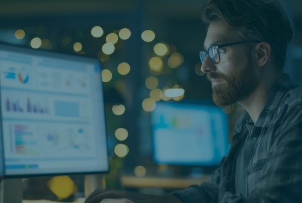 Professional reviewing Security Awareness Training analytics dashboard on dual monitors, analyzing phishing simulation results and employee security performance data in a low-light office setting.